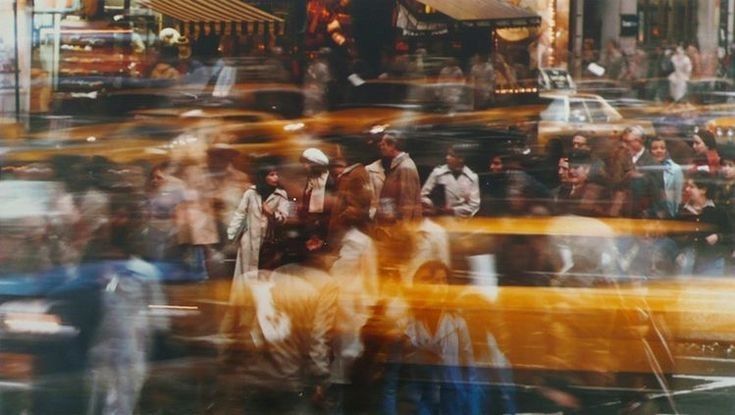 A motion-blurred city street filled with fast-moving yellow taxis and pedestrians crossing, capturing the chaotic pace of urban life.