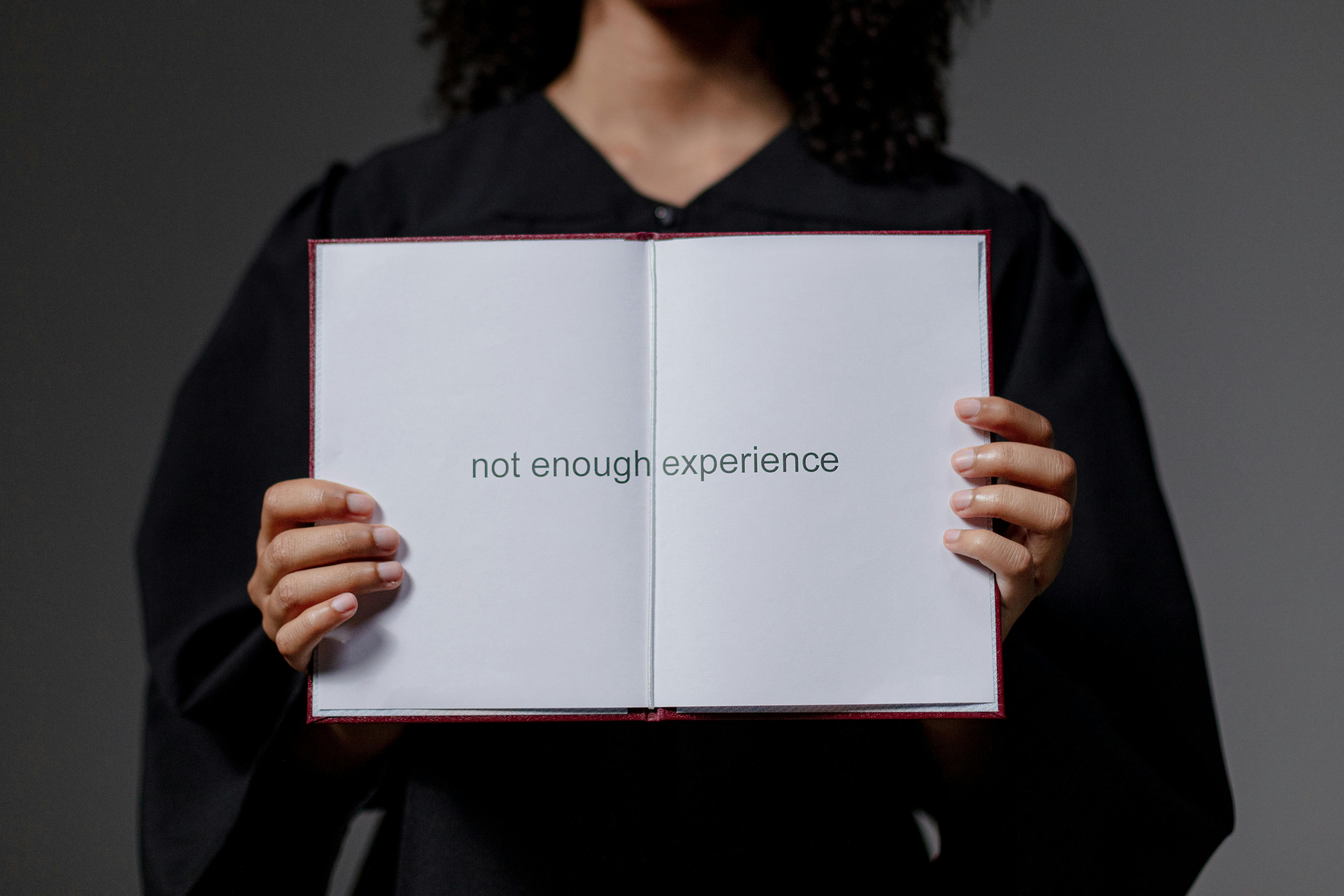 Graduate in academic gown holding open book displaying the words “not enough experience,” symbolizing youth unemployment and entry-level job barriers in India